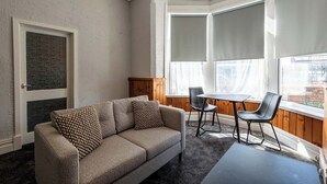 Ein Wohnzimmer mit einer Couch, zwei Sesseln, einem kleinen Tisch und großen Fenstern mit Jalousien.