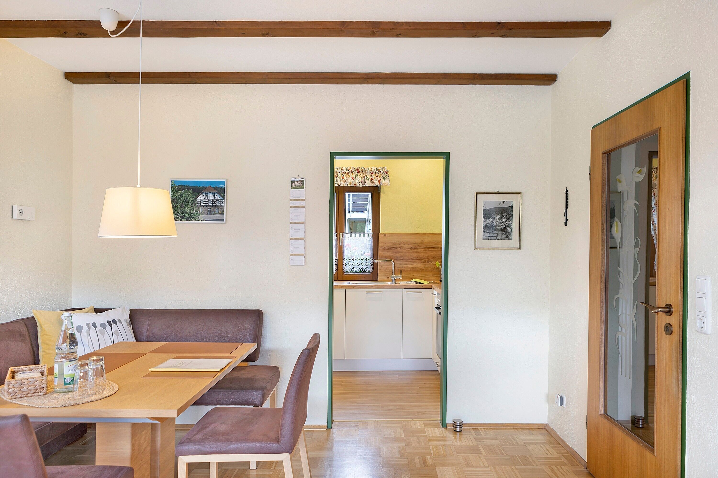 A dining area with a wooden table and chairs, a hanging light fixture, and a view into a kitchen with wooden cabinets and a window.