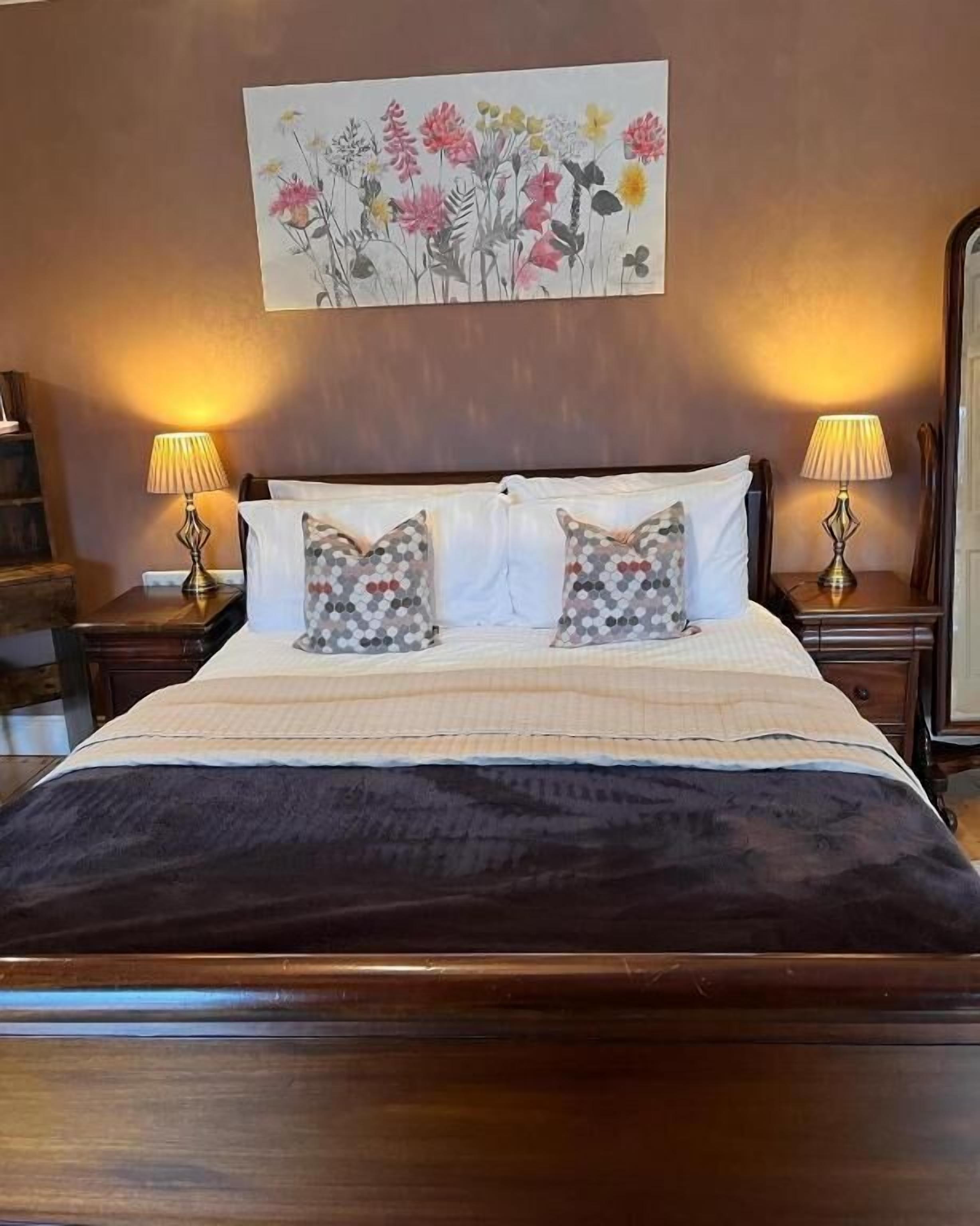 A bedroom with a large bed, two bedside tables with lamps, a painting of flowers on the wall, and a wooden headboard.