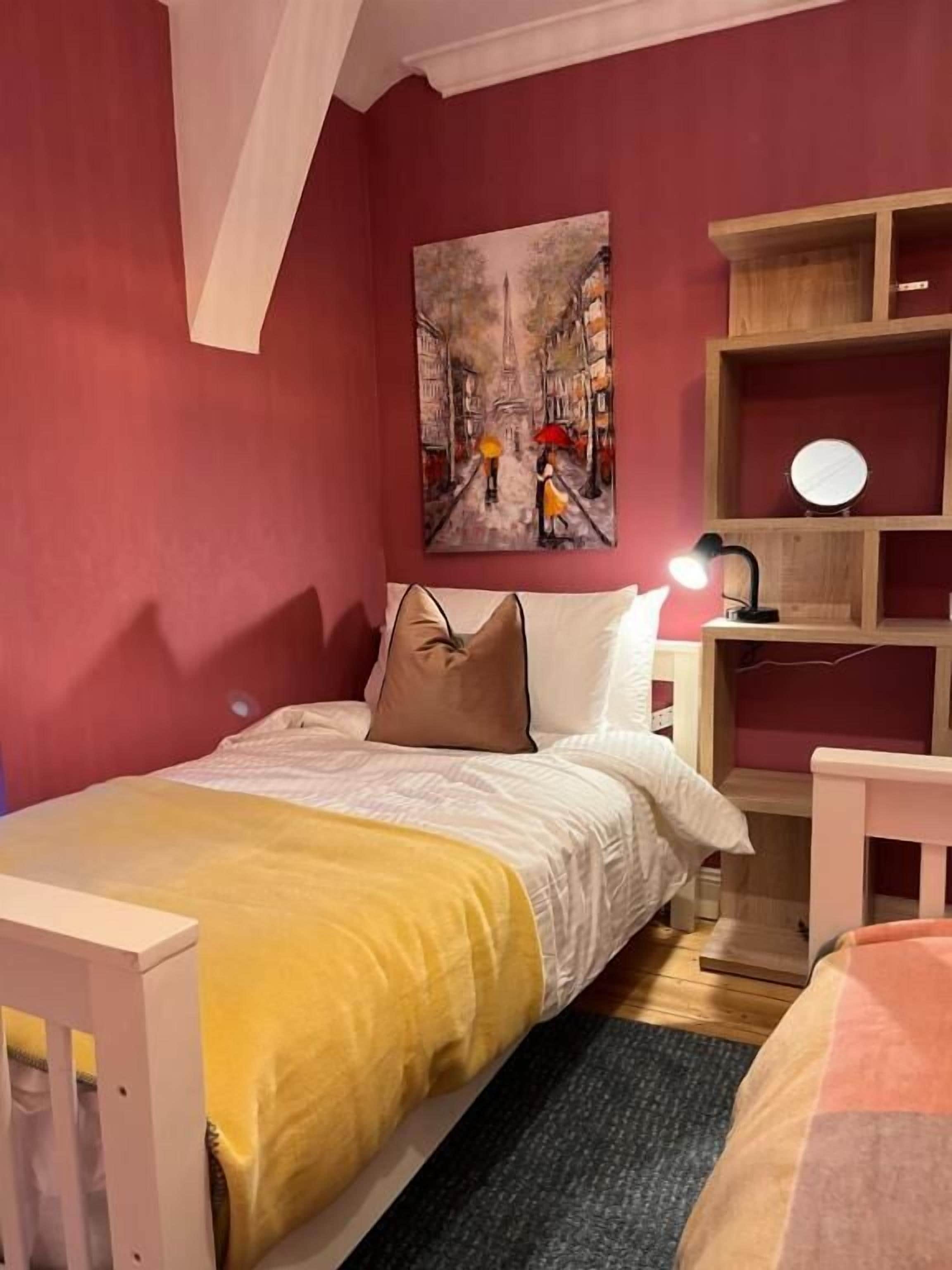 A bedroom with a bed, a nightstand, a bedside table with a lamp, a painting on the wall, and a wooden shelf unit.