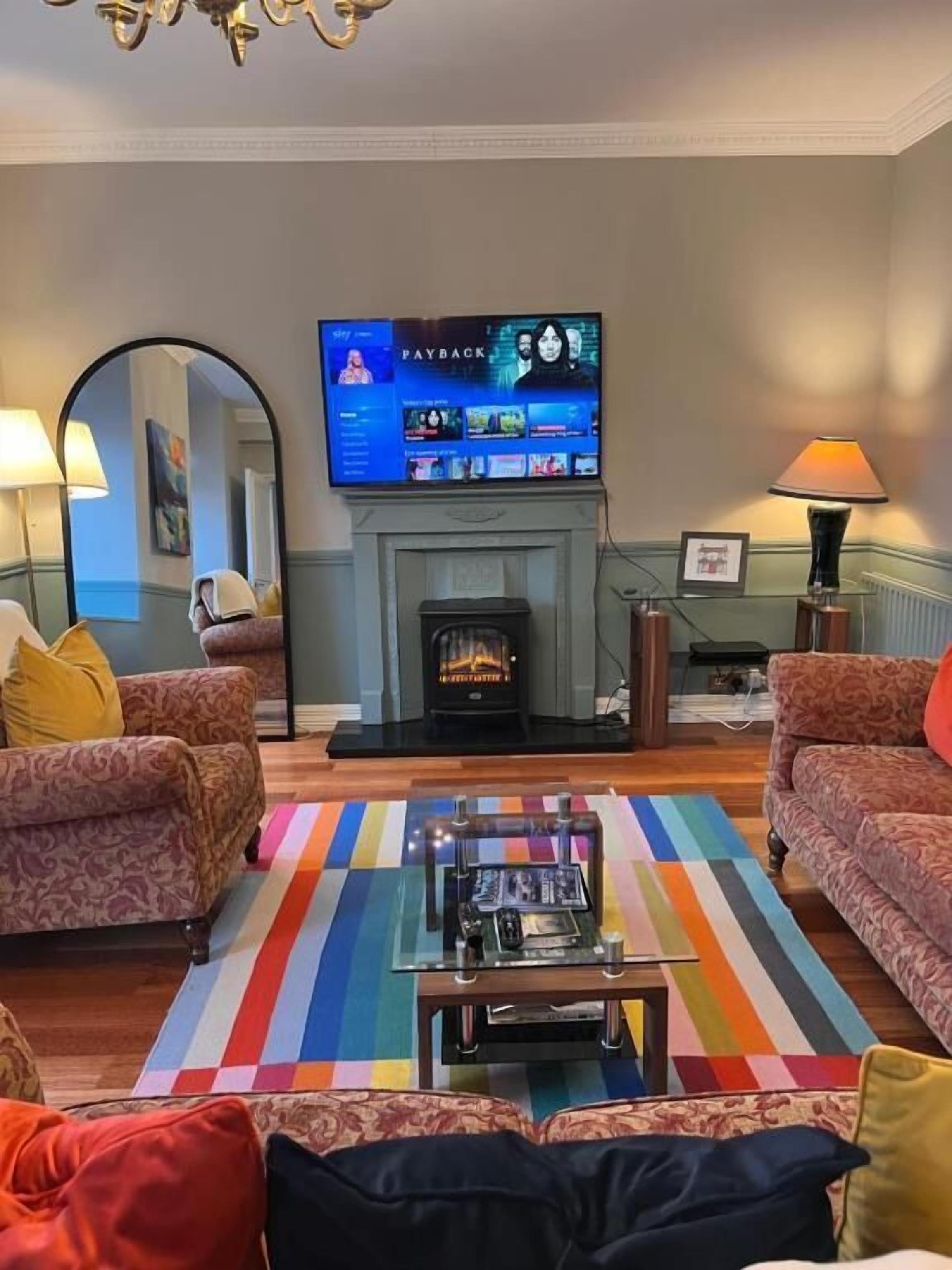 A living room with a fireplace, a television, two patterned sofas, a coffee table, and a colorful rug.