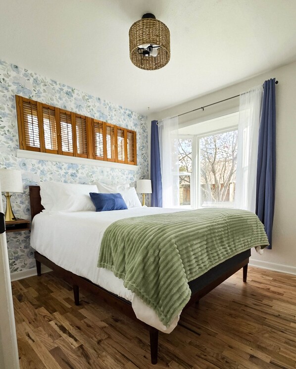 Un dormitorio con una cama, cabecera de madera, una ventana con cortinas, un aplique de pared y un papel tapiz estampado.