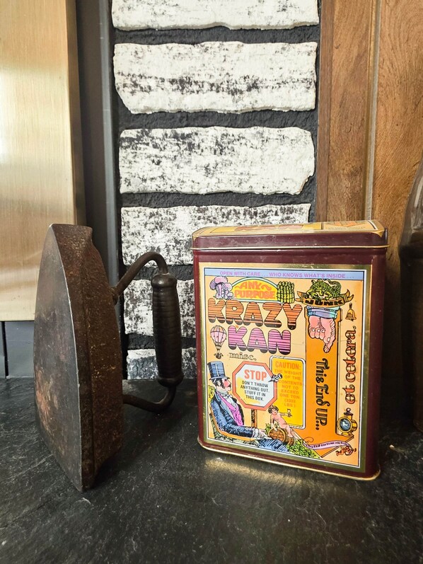 Una lattina vintage etichettata 'KRAZ KAN' con un’etichetta di avvertimento, posizionata su un piano di lavoro nero accanto a un oggetto metallico arrugginito.