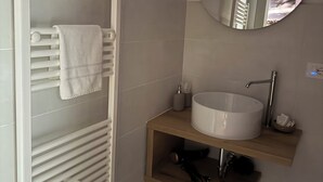 Un baño con toallero, espejo redondo, lavamanos y repisa con artículos de aseo.
