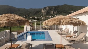 A pool area with lounge chairs and umbrellas, surrounded by mountains and a white building. - Villa Amore-4 Bedroom Villa with Private Pool (Alykes)