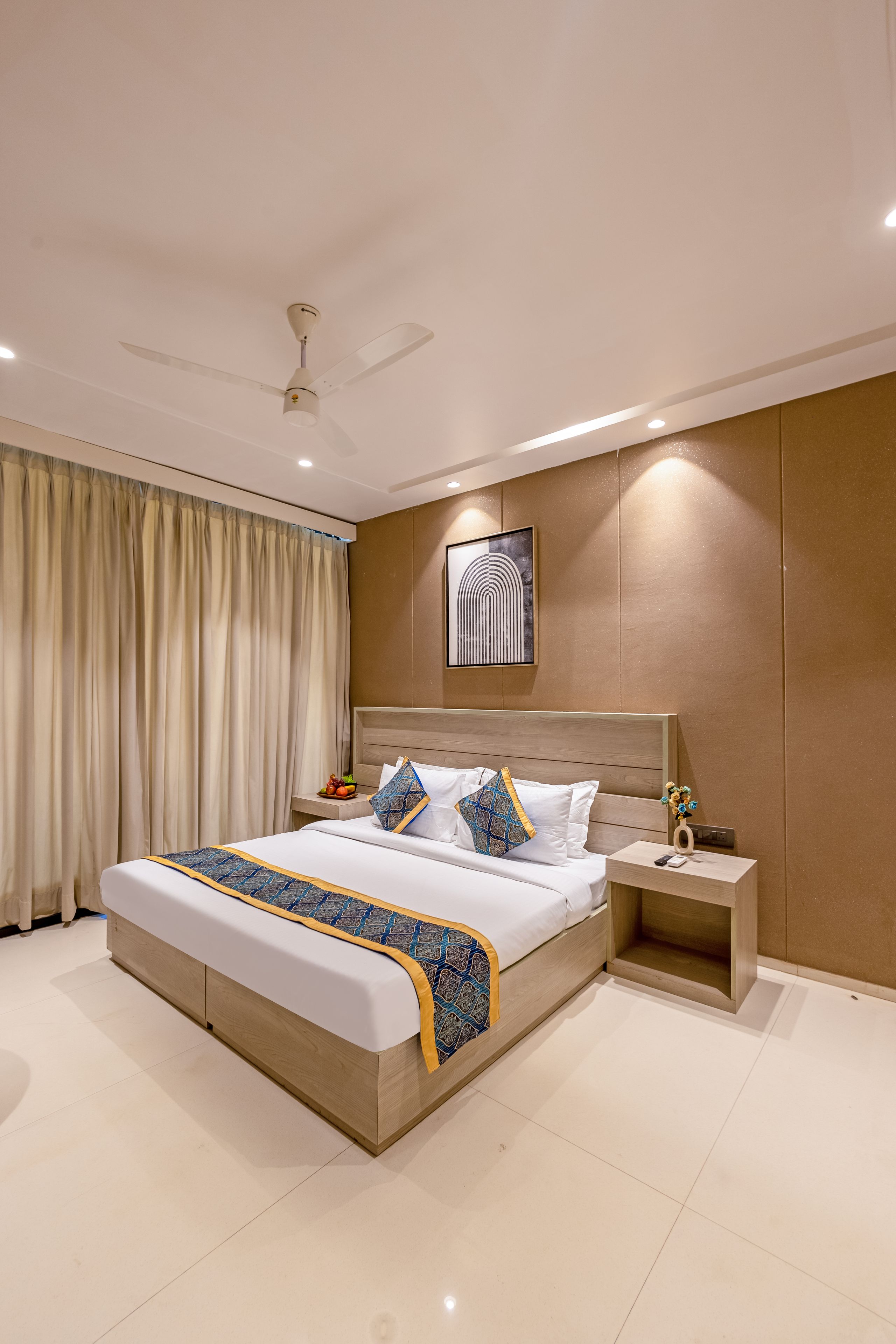 Una habitación de hotel moderna con una cama grande, una mesita de noche, un abanico de techo y una obra de arte colgada en la pared.