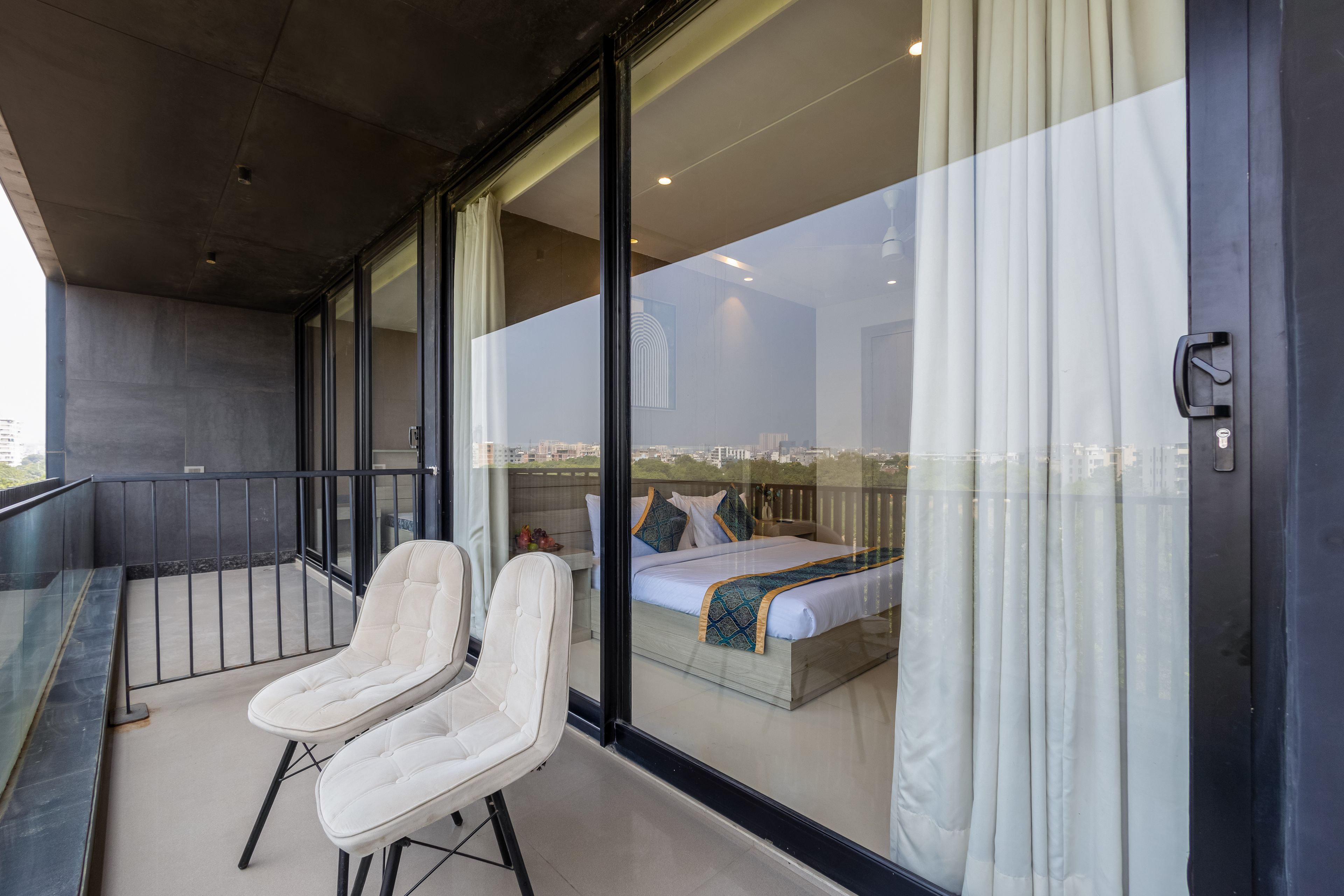 A balcony with two white chairs, a bed with a patterned bedspread, and a city view through large glass doors.