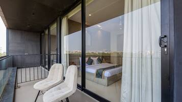 Een balkon met twee witte stoelen, een bed met een beddeken met dessin en een stadszicht door grote glazen deuren.
