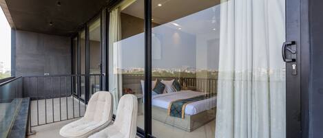 A balcony with two white chairs, a bed with a patterned bedspread, and a city view through large glass doors.