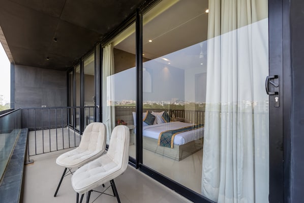 A balcony with two white chairs, a bed with a patterned bedspread, and a city view through large glass doors. - Aurea Avasa (Gurugram)