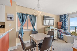 A modern living room with a dining area, a large TV, and a balcony with a view. - Bay Watching the Waves, Large 2 BR with Fitness Center Access in NMB (North Myrtle Beach)