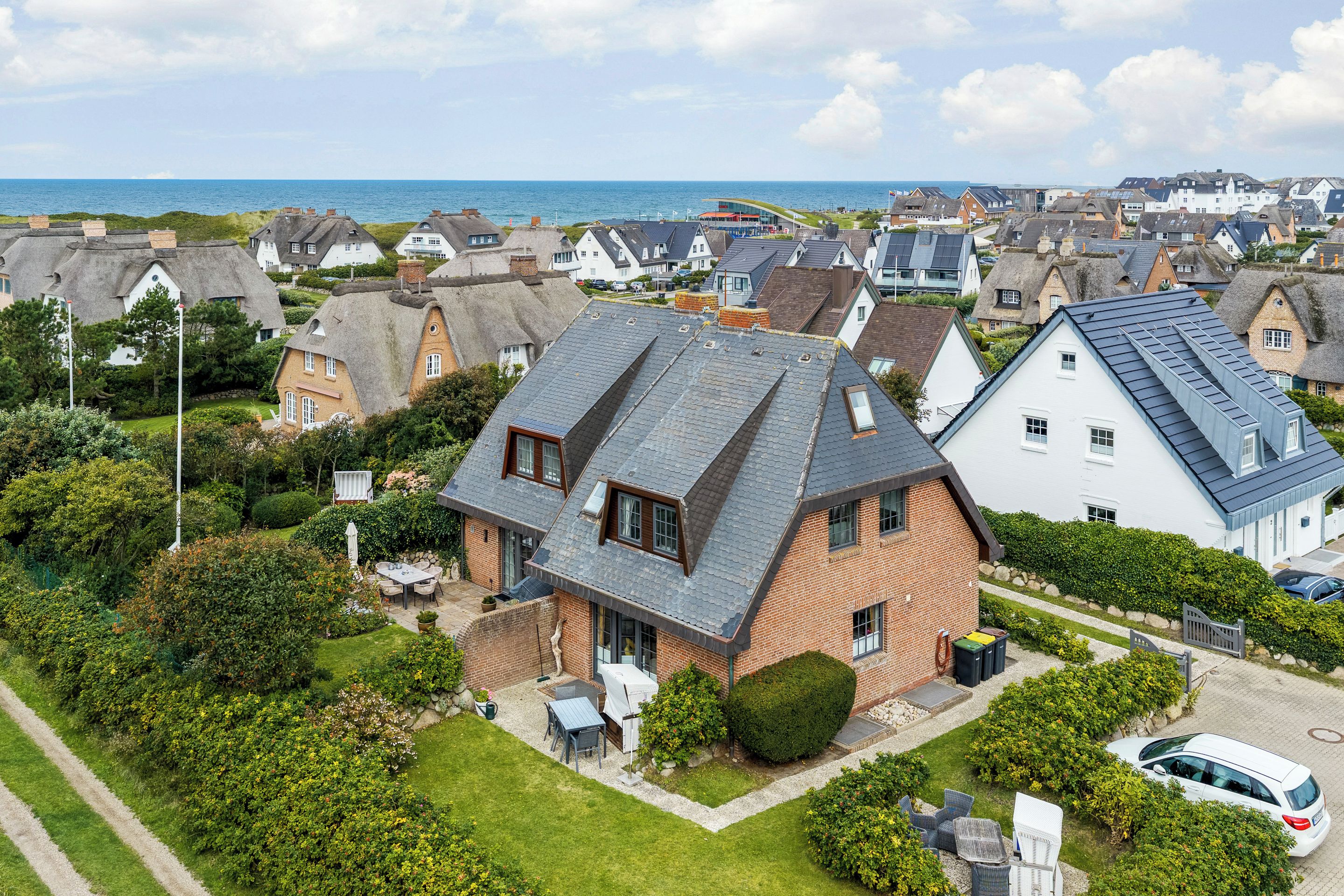 A residential area with a prominent house featuring a sloped roof, a well-maintained garden, and a view of the sea.