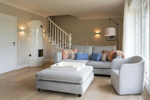 Ein Wohnzimmer mit grauem Sofa, Hocker und Ohrensessel, einer Treppe und einem großen Fenster.
