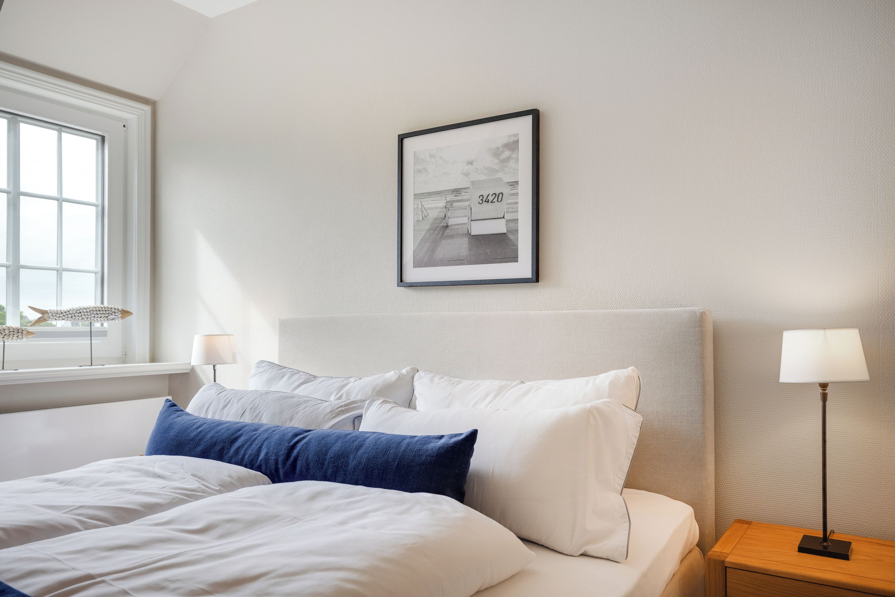 Ein ordentlich bezogenes Bett mit weißer und blauer Bettwäsche, ein gerahmtes Bild an der Wand und eine Lampe auf dem Nachttisch.
