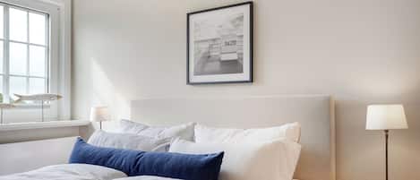 Ein ordentlich bezogenes Bett mit weißer und blauer Bettwäsche, ein gerahmtes Bild an der Wand und eine Lampe auf dem Nachttisch.