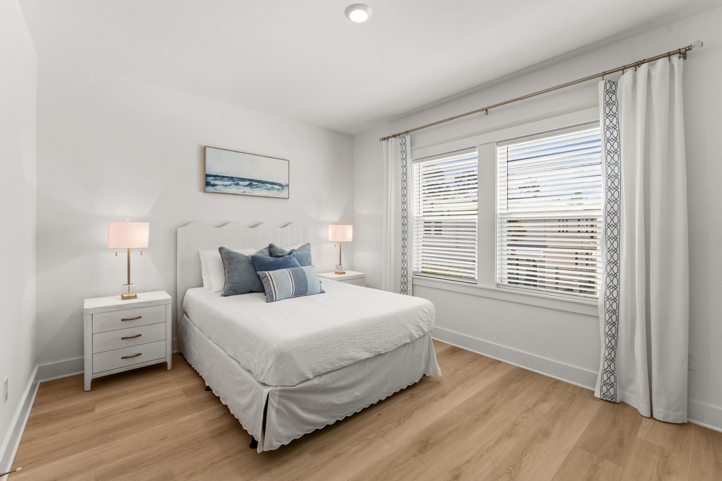 A bedroom with a bed, bedside tables, lamps, a window with blinds, and a painting on the wall.