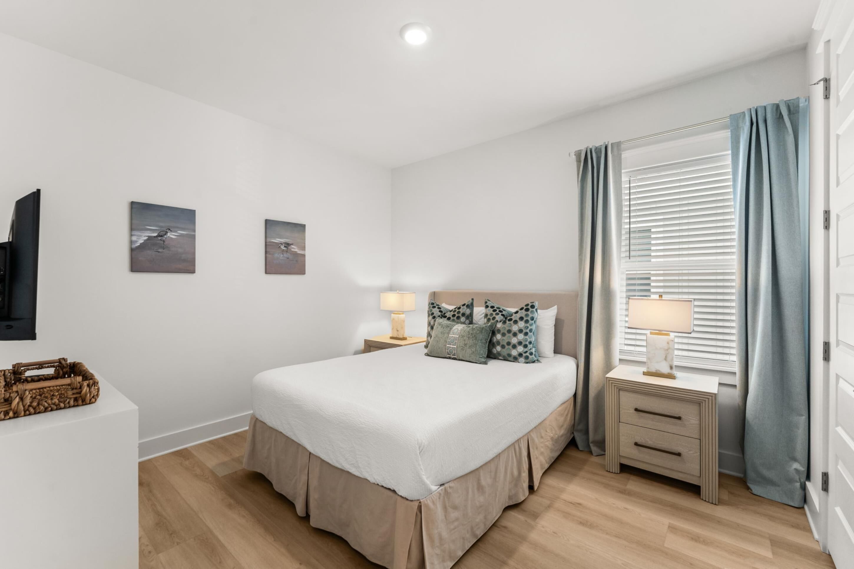 Une chambre à coucher avec un lit, des tables de chevet, une télévision, des œuvres d’art et une fenêtre avec des rideaux.