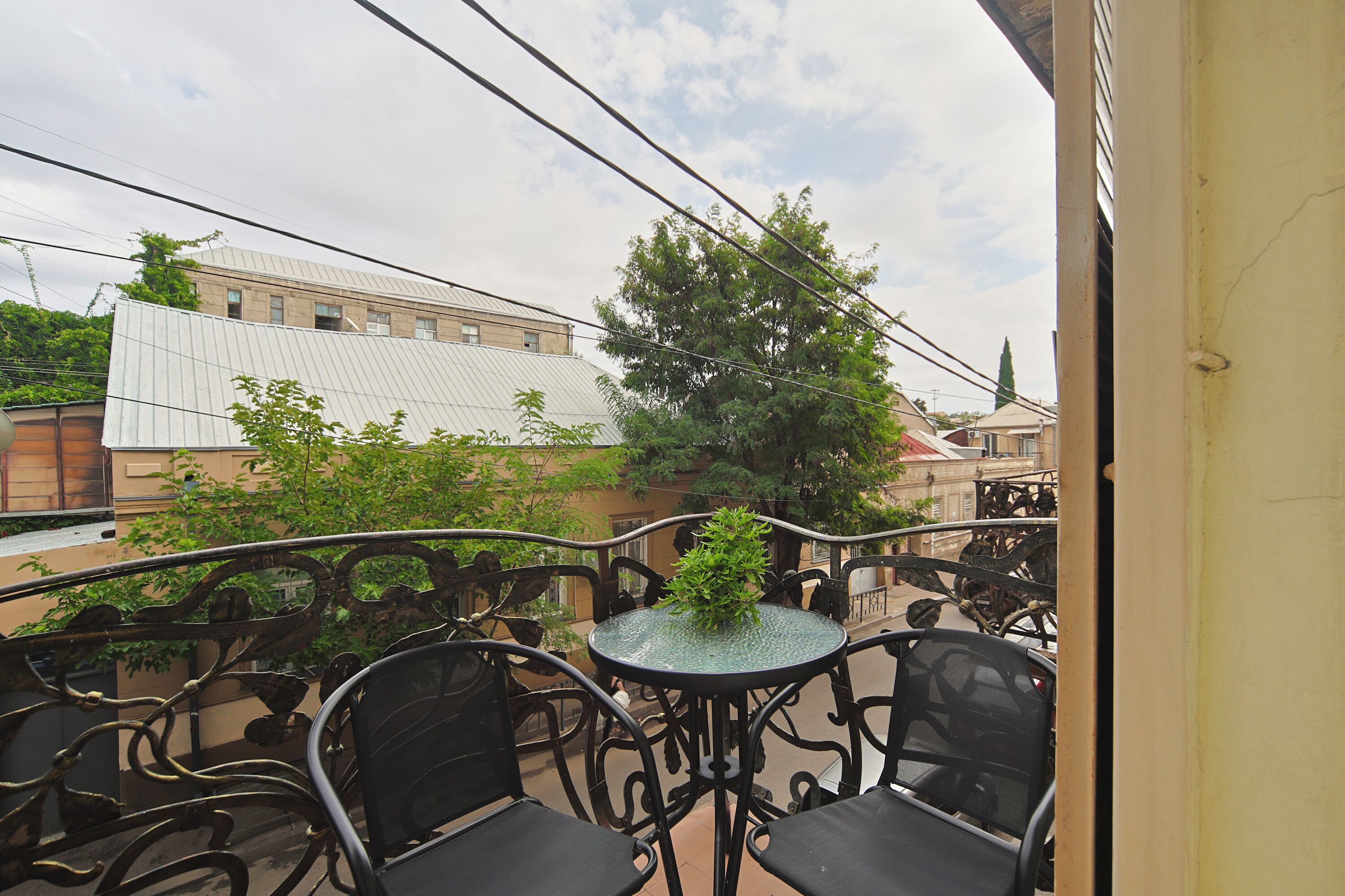 A balcony with metal chairs and a table, overlooking a building with a white roof and trees.