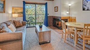 Sala de estar com sofá, mesa de centro e mesa de jantar. Há uma lareira e uma vista através de uma grande janela.