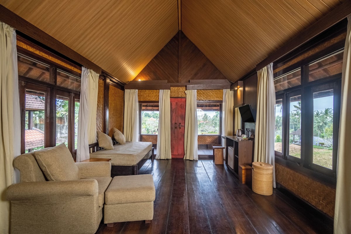 A room with a wooden ceiling, large windows, a bed, a sofa, a TV, and a small table.