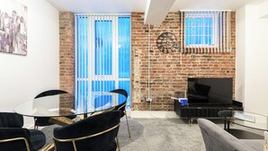 Un salon moderne avec une table en verre, des chaises noires, un canapé gris, un téléviseur et un mur en briques.
