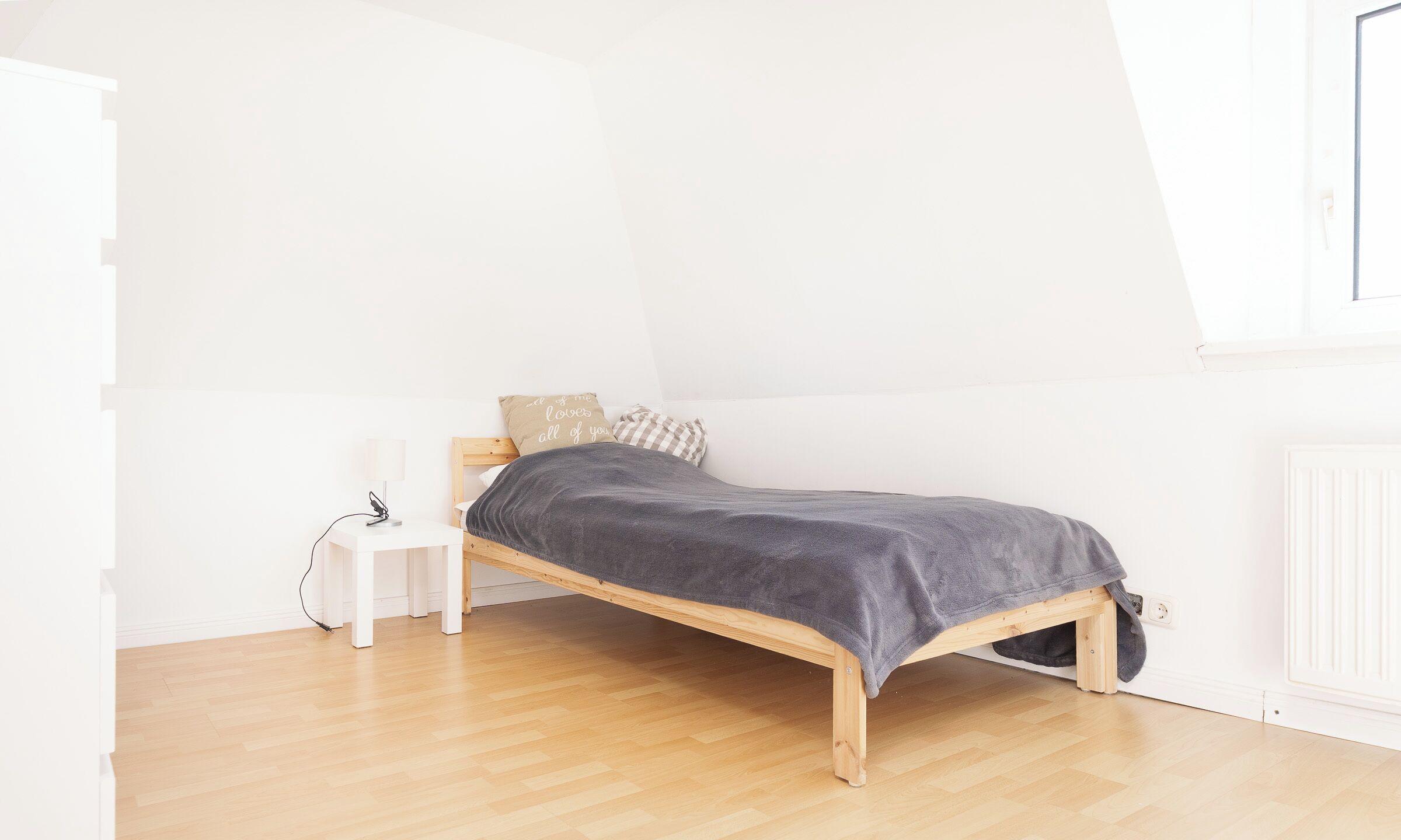 Een kleine eenpersoonskamer met een houten bedframe, een grijze dekbedovertrek, een witte nachtkastje met een lamp en een raam met uitzicht op de lucht.