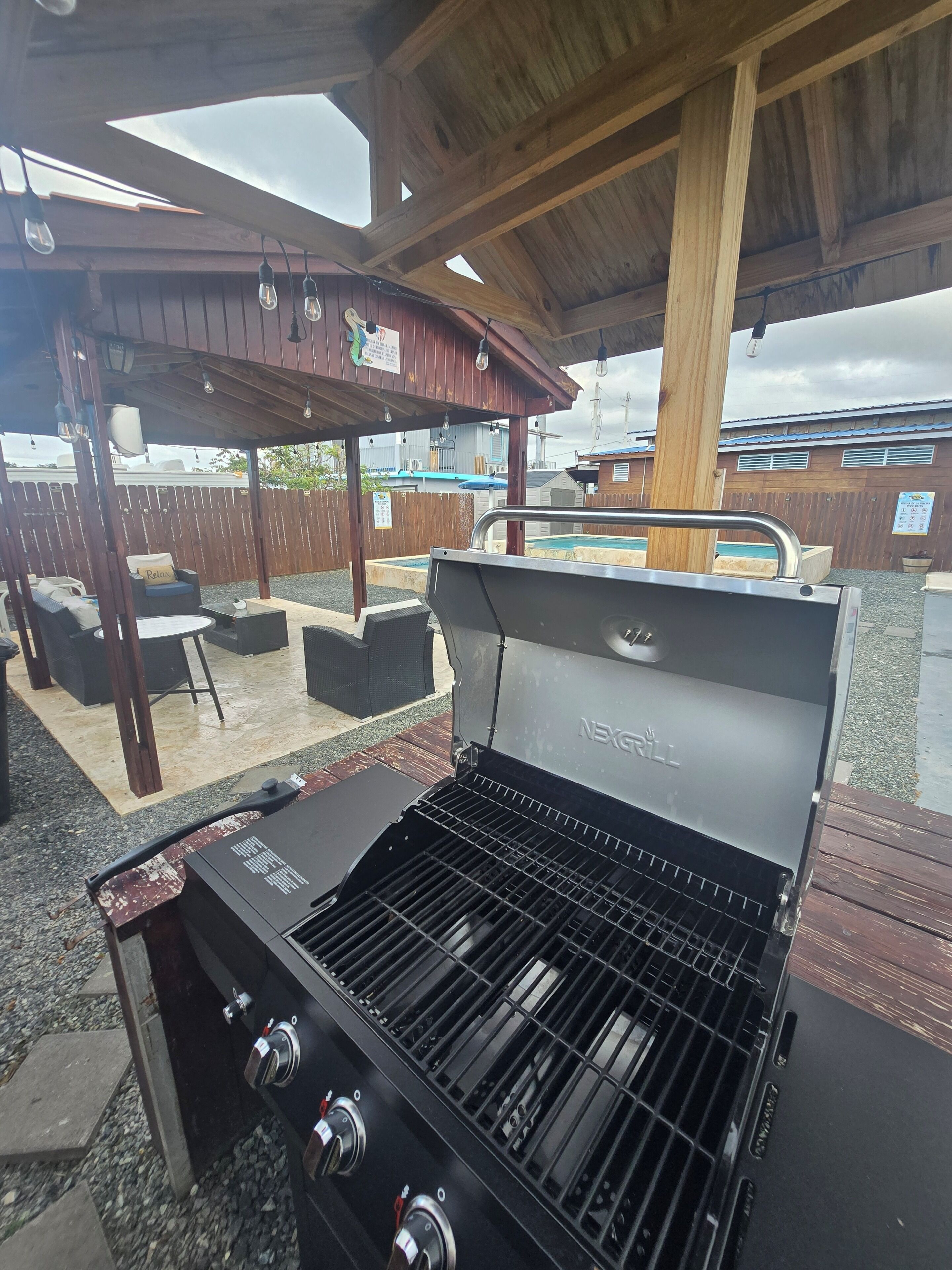 Ein Grillbereich im Freien mit einer hölzernen Pergola, einem Pool im Hintergrund und einer umzäunten Terrasse.