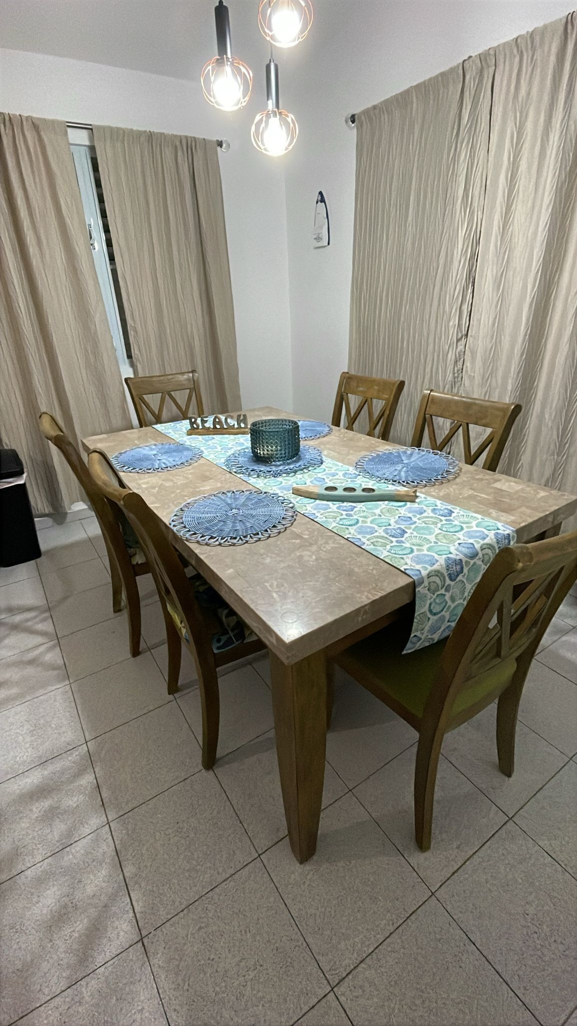 A dining table with a patterned tablecloth, surrounded by wooden chairs, in a room with curtains and a light fixture.