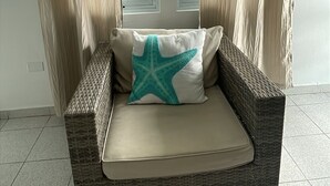 Um sofá de vime com almofada e um travesseiro em formato de estrela-do-mar, posicionado em frente a uma janela com cortinas bege.