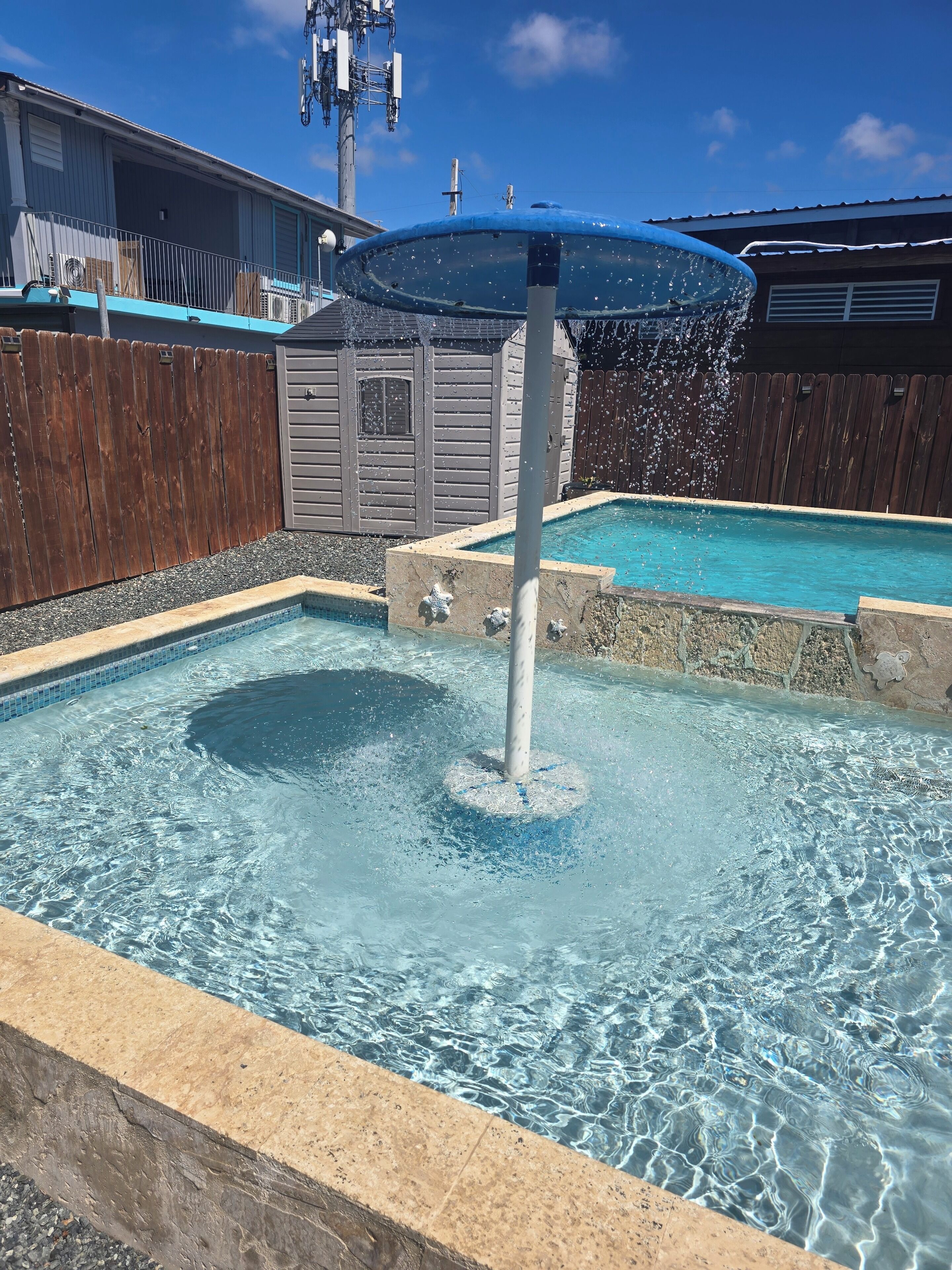Una piscina con fuente, rodeada por una valla de madera y un edificio de fachada gris.