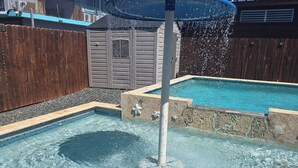 Uma piscina com uma fonte, rodeada por uma cerca de madeira e um edifício com fachada cinzenta.