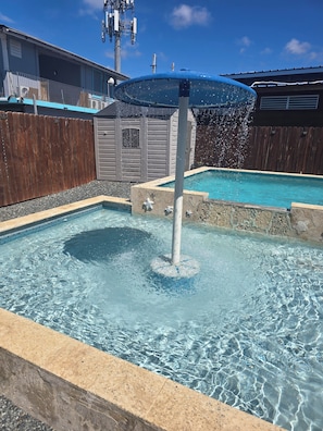 Una piscina con fuente, rodeada por una valla de madera y un edificio de fachada gris.