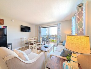 Una sala de estar con sofá, una mesa de comedor con sillas, un televisor montado en la pared, un balcón con vistas a la ciudad y una lámpara sobre una mesita auxiliar.