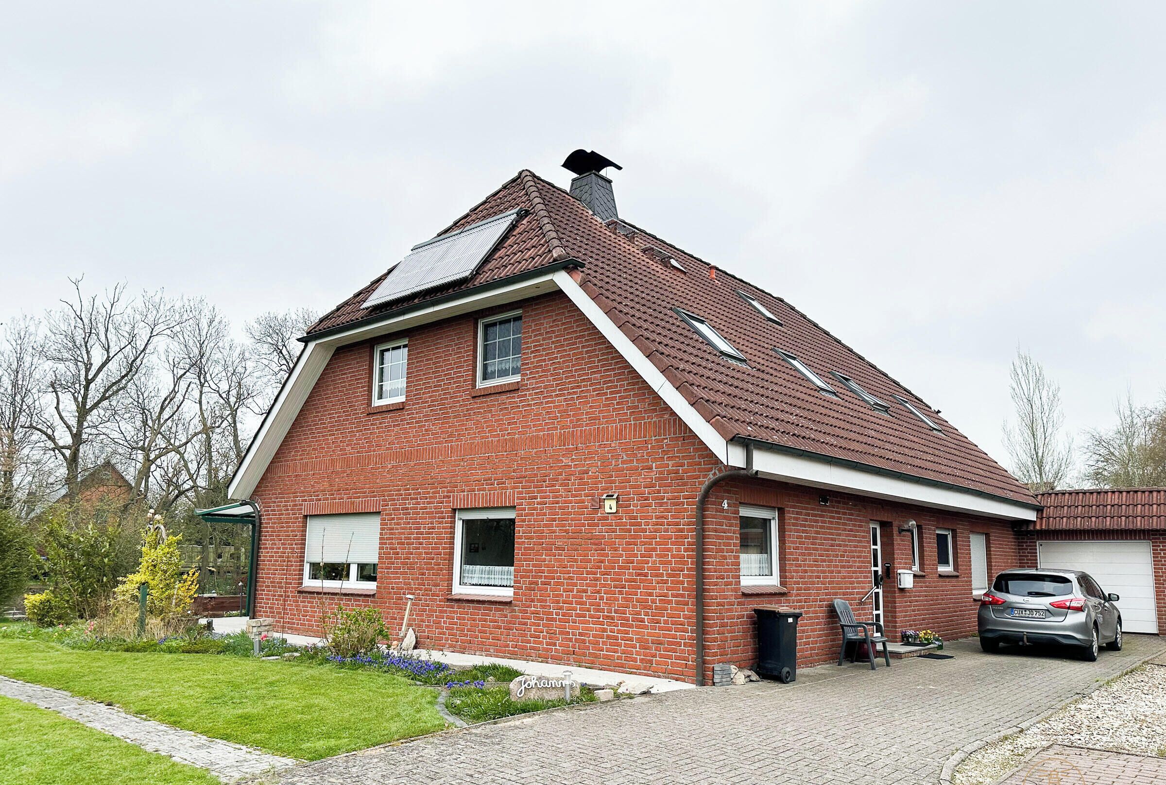 Ein rotes Backsteinhaus mit geneigtem Dach, ein Auto auf der Auffahrt und ein Garten mit Blumen und Sträuchern.