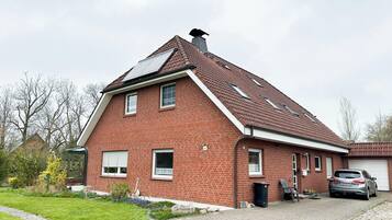 Ein rotes Backsteinhaus mit geneigtem Dach, ein Auto auf der Auffahrt und ein Garten mit Blumen und Sträuchern.