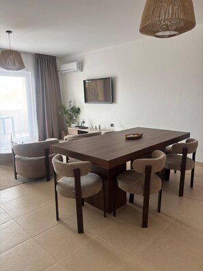 A modern dining area with a large wooden table, six upholstered chairs, a television mounted on the wall, and a large window with curtains. - Casa Tortuga Sal Apartment 1st Floor With Balcony (Sal)