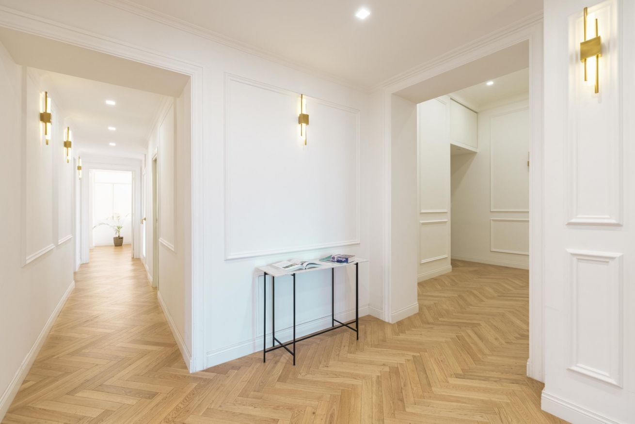 A hallway with a herringbone floor, white walls, and a table with magazines.