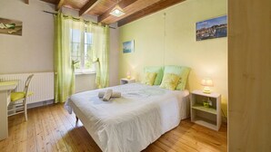 A bedroom with a wooden floor, a bed with green pillows, a nightstand with a lamp, a chair, and a window with curtains.