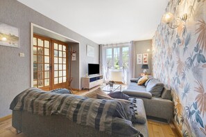 A living room with a sofa, a television, a coffee table, and a window with curtains. - Holiday Home 'Gîte De L'arthour' with Private Garden and Wi-Fi (Notre-Dame-de-Cenilly)