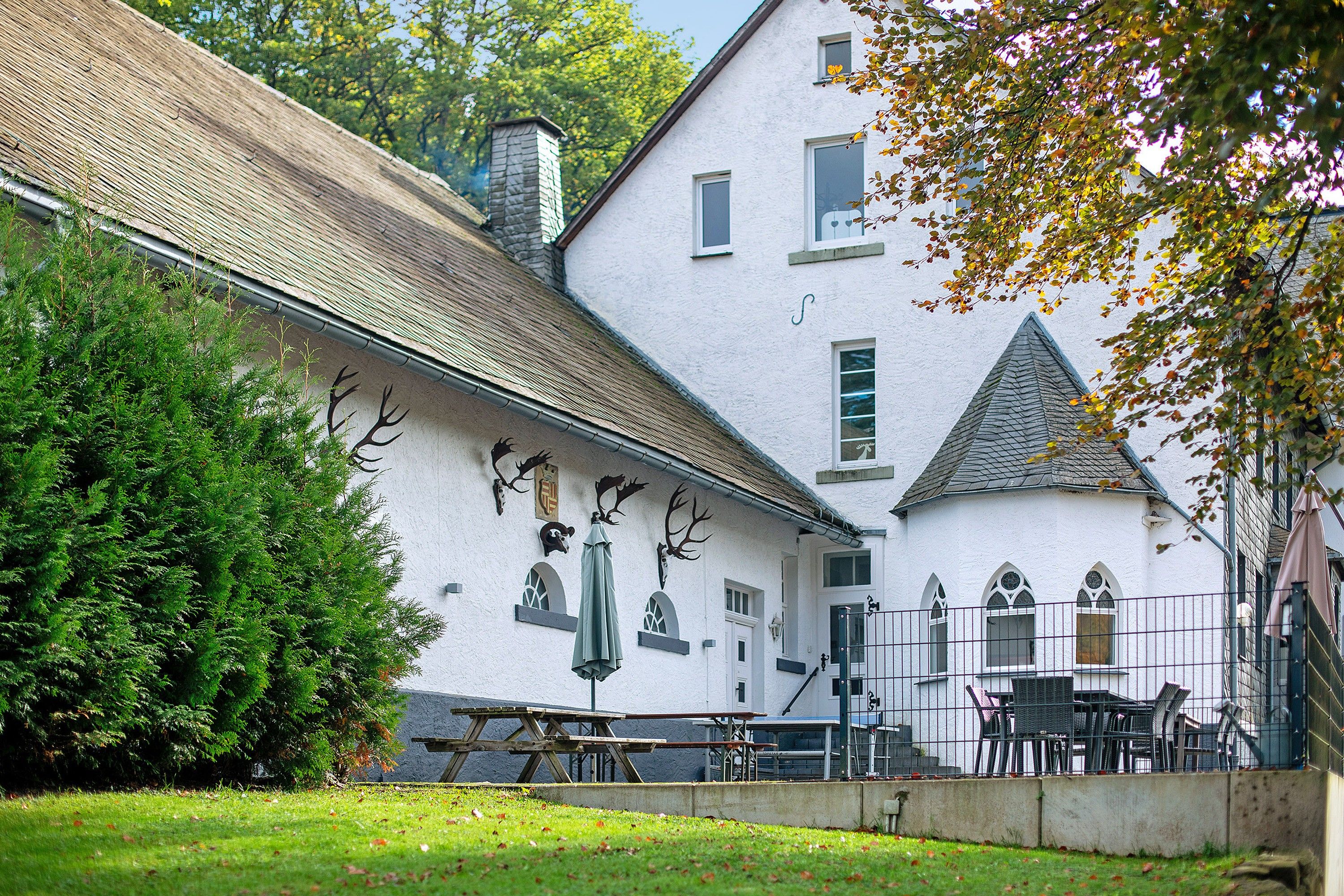 A white building with a steeply pitched roof, a stone chimney, and a series of animal heads mounted on the wall.