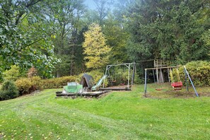 Un terrain de jeux avec des balançoires et un toboggan, entouré d’arbres et d’herbe.