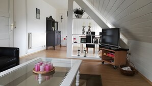 Ett vardagsrum med ett soffbord i glas, en svart soffa, en tv på ett tv-bord, en bokhylla och en trappa.