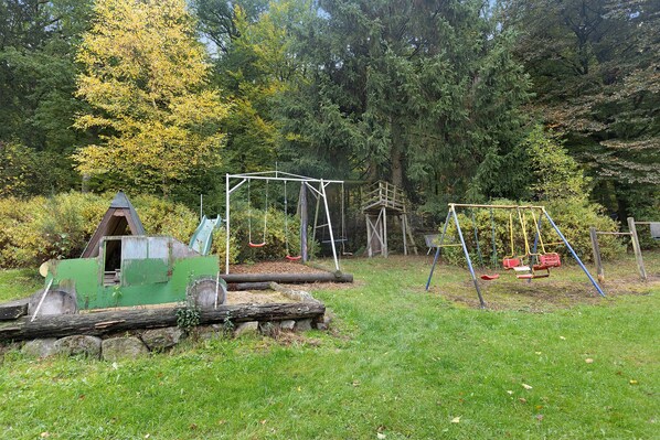 Un parque infantil con columpios y un tobogán, rodeado de árboles y césped.