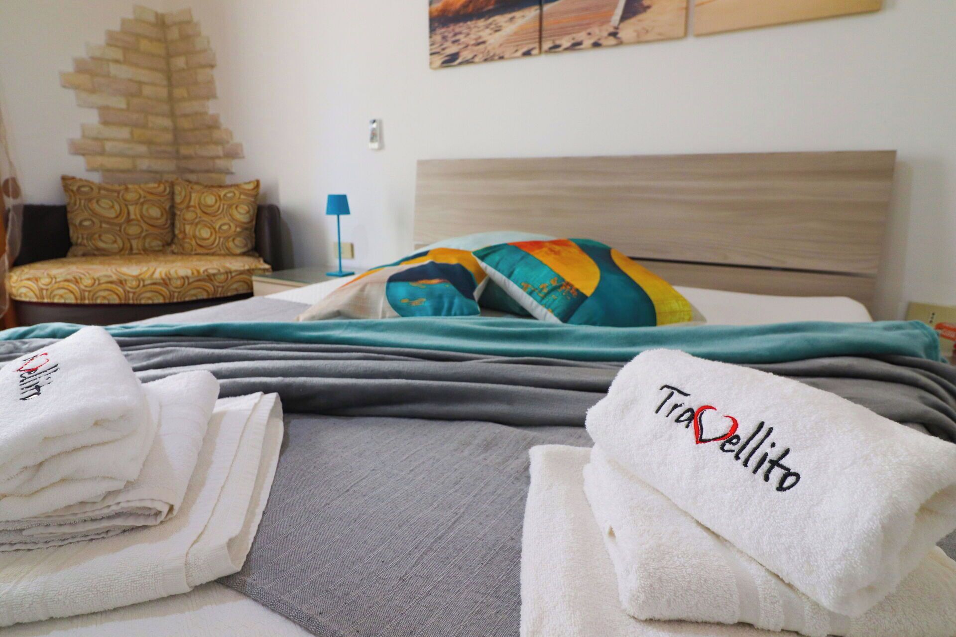 A neatly made bed with a grey and teal blanket, a colorful pillow, and a stack of white towels with 'Tranquillo' embroidered on them.