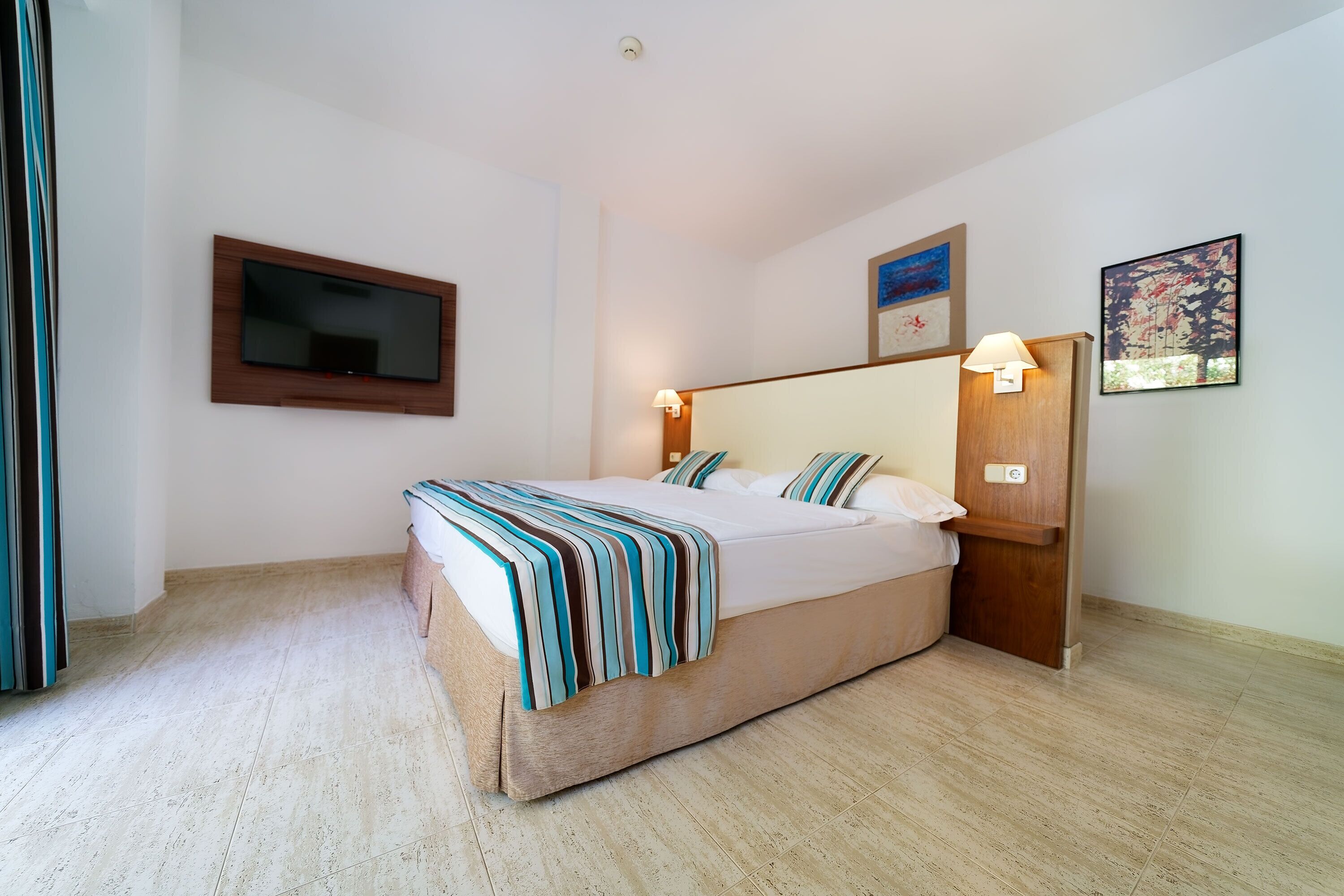 Een slaapkamer met een bed, een tv aan de muur, een nachtkastje met een lamp en een schilderij.