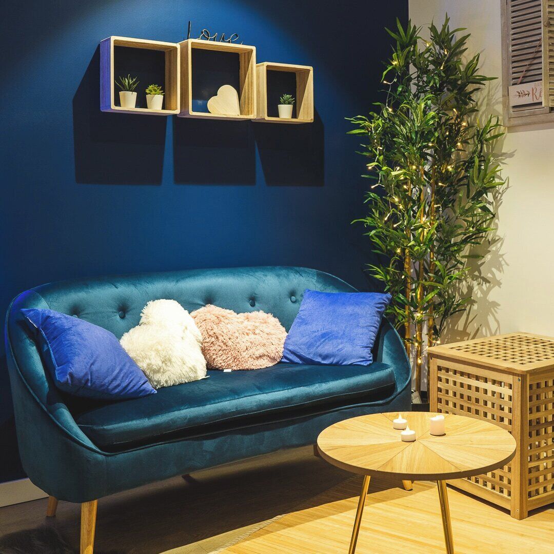 Ein blaues Sofa mit Kissen, ein kleiner runder Holztisch mit Kerzen und eine hohe Pflanze mit Lichtern.