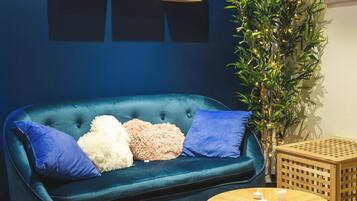Ein blaues Sofa mit Kissen, ein kleiner runder Holztisch mit Kerzen und eine hohe Pflanze mit Lichtern.
