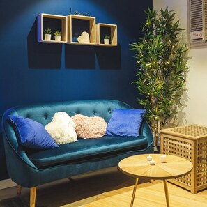 Un canapé bleu avec des coussins, une petite table ronde en bois avec des bougies et une grande plante décorée de guirlandes lumineuses.