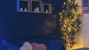 Un sofá azul oscuro con dos cojines, una pequeña mesa redonda de madera con velas, y una planta de bambú con luces.