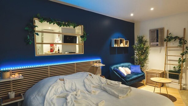 Un dormitorio moderno con cama, un sillón azul, una mesita de noche, un espejo y una planta pequeña.